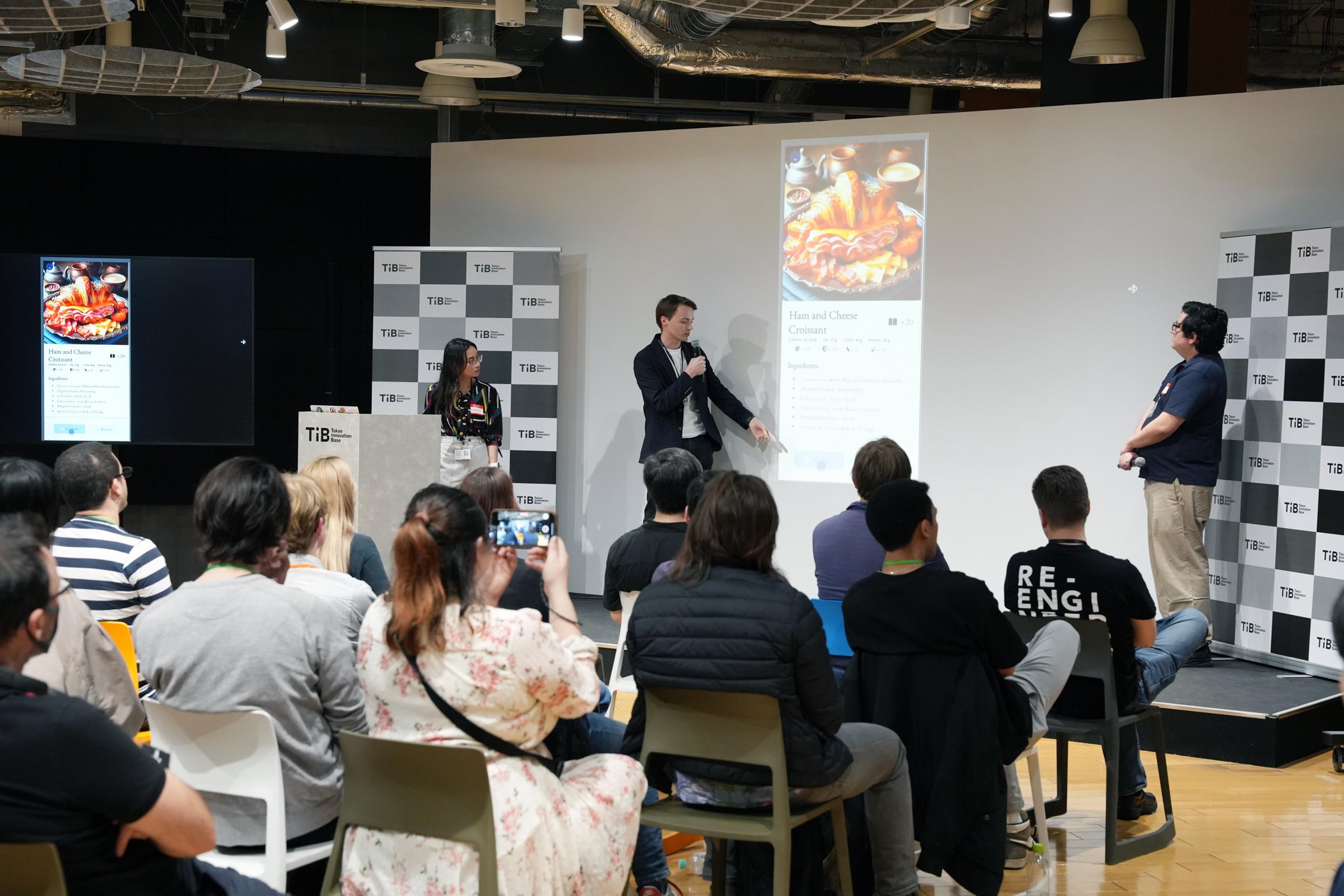 Matt presenting a full-stack app to a seated audience at Tokyo Innovation Base, a nutrition screen for a ham and cheese croissant projected on the wall behind him