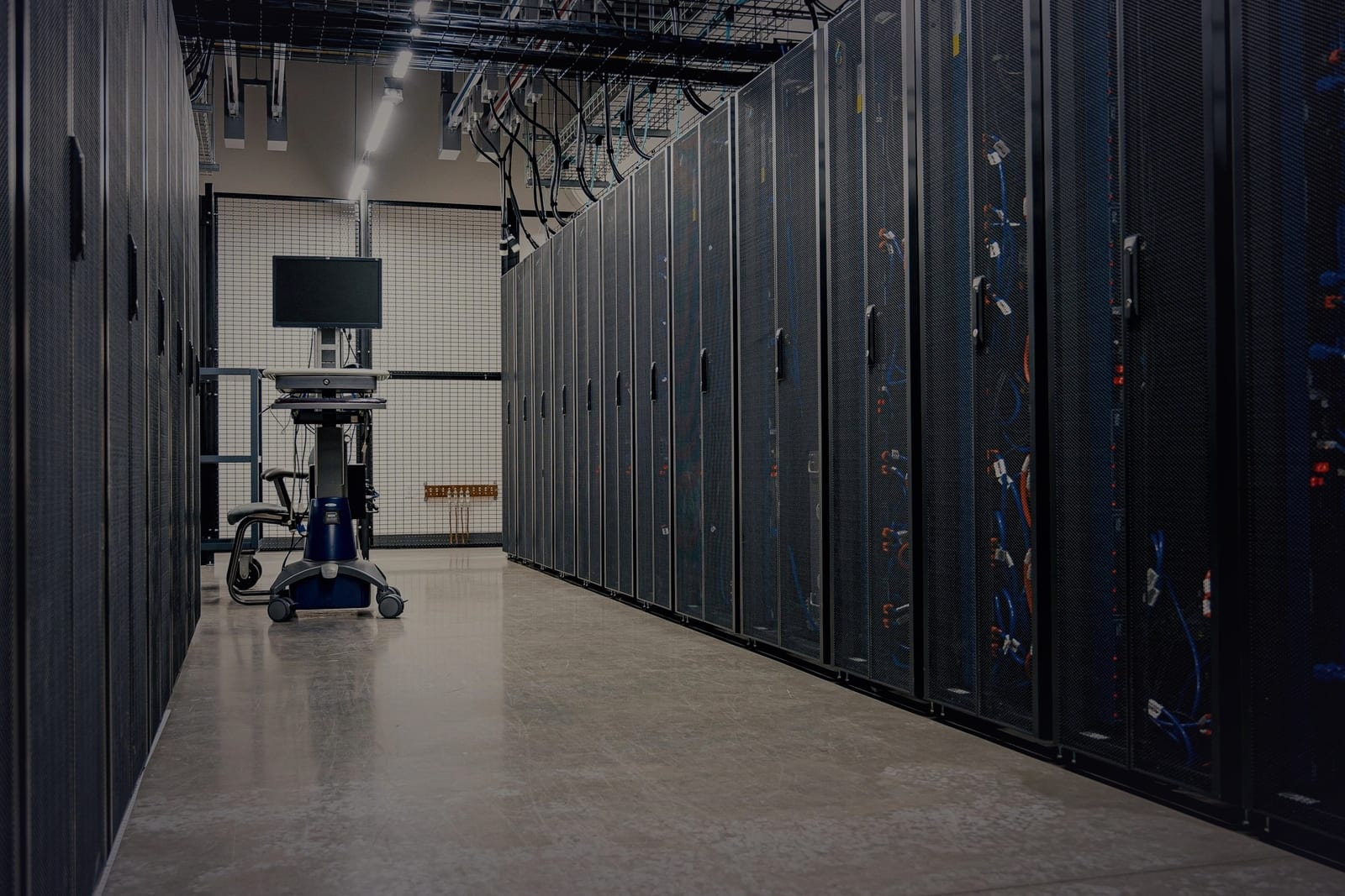 A dimly lit data center aisle lined with server racks, with a single unattended terminal on a cart at the far end